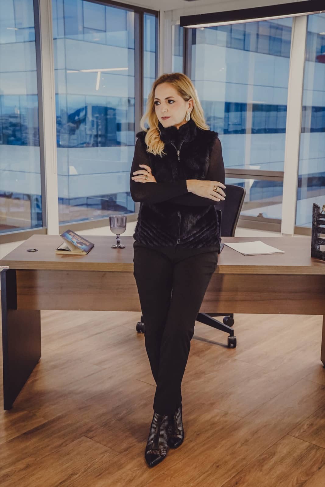 Paty Ivo de braços cruzados, encostada na mesa do escritório, olhando pela janela